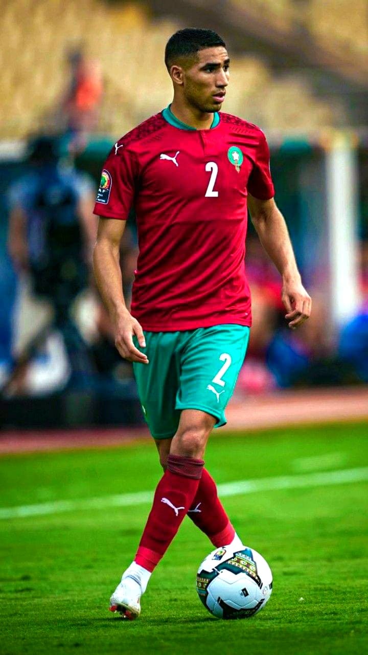 A soccer player in a red and green Morocco jersey with number 2 controls a ball on the field during a match, as fans around the world follow the action live through IPTV Italy.